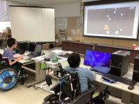 電動車いすの２名の生徒がスクリーンを見ながらシューティングゲームをやっている