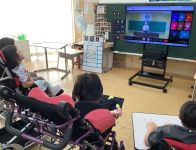 教室で、数人の児童たちが大きなモニターの前に座り、校長先生の話を聞く様子。