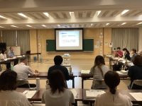 中央にいる男性が話しているようです。壁にはプロジェクターのスクリーンがあり、スクリーンにはスライドが映し出されています。