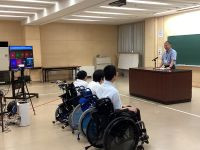 校長先生の話を３人の生徒が聞いている。後方にリモート用のモニターやカメラが設置されている。