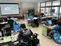 車椅子に座った数名の生徒が、教室でデスクトップパソコンを使って作業をしている。スクリーンに建物の3Dモデルが写っている。