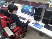 電動車いすに座った生徒がデスクトップパソコンでタッチパッド付きキーボードとマウスを使って作業をしている。モニターにはUnityの作業画面が写っている。