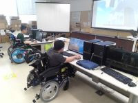 車椅子に座った2名がパソコンで作業する教室の様子。奥のスクリーンには建物の3Dモデルが映し出されている。