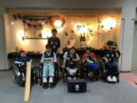 ハロウィン装飾が施された会場で、ボッチャ大会参加者と見られる6人が並んで写る集合写真。車椅子の5名と立っている1名は黒いユニフォーム姿で、うち1名は「郡山支援B」のボードを持っている。左端にボッチャ用のスロープ、中央にはボールバッグが置かれている。