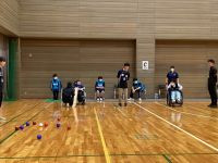 体育館でボッチャの試合を行っている様子の写真。車椅子利用者を含む9名が、木の床に引かれたコート上にいる。中央付近には、赤いボールと青いボール、白いジャックボールが散らばっている。コート奥の壁には「C」と書かれた白いプレートが見える。参加者はマスクを着用している。