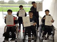 陸上競技場で、車椅子に乗った5名の若者が、賞状とメダルを持って並んでいる写真。全員が笑顔を見せ、誇らしげな表情をしている。背景には屋外のトラックや芝生のフィールドが見え、屋根付きの観客席下で撮影された様子。賞状には「賞 陸上競技」の文字が確認できる。