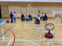 体育館で、車いすに乗っている女の子が、フロアの点数やリングに向かってビーンバッグ（お手玉）を投げている。周りでは他の参加者が見守っている。