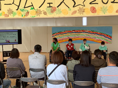 校内で開かれた学部行事公開「オータム☆フェス２０２５」の発表の様子。チームブルースカイの４人の生徒たちが観客に向けてボッチャの説明をしている。背景には虹の絵、左手には「どういう人が参加できるか」「ボッチャ」などが表示されたモニターがある。頭上には「オータムフェス☆２０２５」と部分的に読める横断幕が掲げられている。