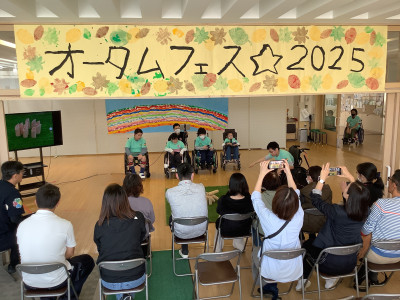 校内で開かれた学部行事公開「オータム☆フェス２０２５」の発表の様子。「からふるグループ」の６人の生徒たちが観客に向けてモルックについて発表し、１人の生徒がモルックの実演をしている。背景には虹の絵、左手にはモルックの実演の様子を映したモニターがある。頭上には「オータムフェス☆２０２５」と書かれた黄色の横断幕が掲げられている。