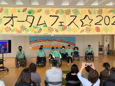 校内で開かれた学部行事公開「オータム☆フェス２０２５」の発表の様子。１０人の生徒が並び、客席では観客がスマートフォンを向けて撮影している。背景には虹の絵、頭上には「オータムフェス☆２０２」と部分的に書かれた黄色い横断幕が掲げられている。