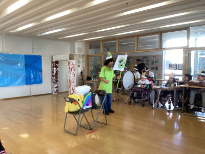 会場の自立活動室で、音楽劇「十二支のおはなし」を発表している様子。中央には、蛇のお面をかぶり緑のTシャツを着た生徒が立っている。手には龍が印刷された紙を持っている。会場右手側には、十二支の動物のお面をかぶった生徒が映っている。近くには、大太鼓やギターがあり、劇の動きに合わせて効果音を演奏している。