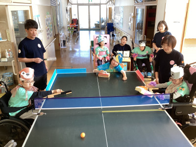動物の被り物をした車椅子の生徒が卓球バレーをしている様子。中央の生徒は笑顔で楽しそうな表情。卓球台には青いネットが使われ、奥の廊下には車椅子などが置かれている。奥の得点板には「1」の文字が見える。