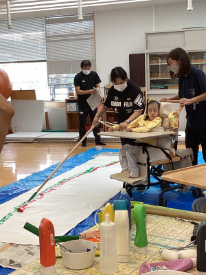 室内で、車椅子に座った生徒が、劇のバック画を制作している。床に敷かれた青いシートと長い白い紙の上で、長い棒に付けたローラーを使って虹の絵を描いている様子。
