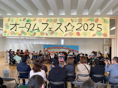 会場の自立活動室で、観客が、車椅子に座って衣装を身に着けた生徒たちが行う劇「オズの魔法使い」を見ている。劇の最後に生徒が横一列になり、紐につながった鈴を持ちながら歌を歌っている様子。 上部には「オータムフェス☆２０２５」と書かれ、紅葉のスタンプで飾られた横断幕が掲げられている。