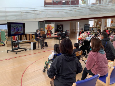 ① プレイルームでの講話の様子。前方では、マスクを着用した学校歯科医がモニターを見ながら話している。後方には、車椅子や椅子に座り保護者と子どもたち、先生が聴講している。