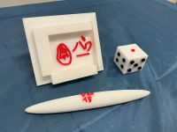 青い布の上に、白い3Dプリント作品が3つ並んでいる。左は立てかけられた額のような形で、中央に赤い文字とマークが立体的に配置されている。中央奥には白いサイコロがあり、黒と赤で目がついている。手前には白い円筒状の細長いオブジェが置かれ、赤い文字が刻まれている。