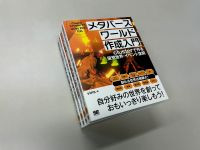 机の上に『メタバースワールド作成入門』という書籍が４冊積まれている。表紙には仮想空間でアバターが炎のような光に照らされたイラストが描かれており、『clusterで作る仮想世界・イベント空間』『自分好みの世界を創っておもいっきり楽しもう！』などの文字が印刷されている。